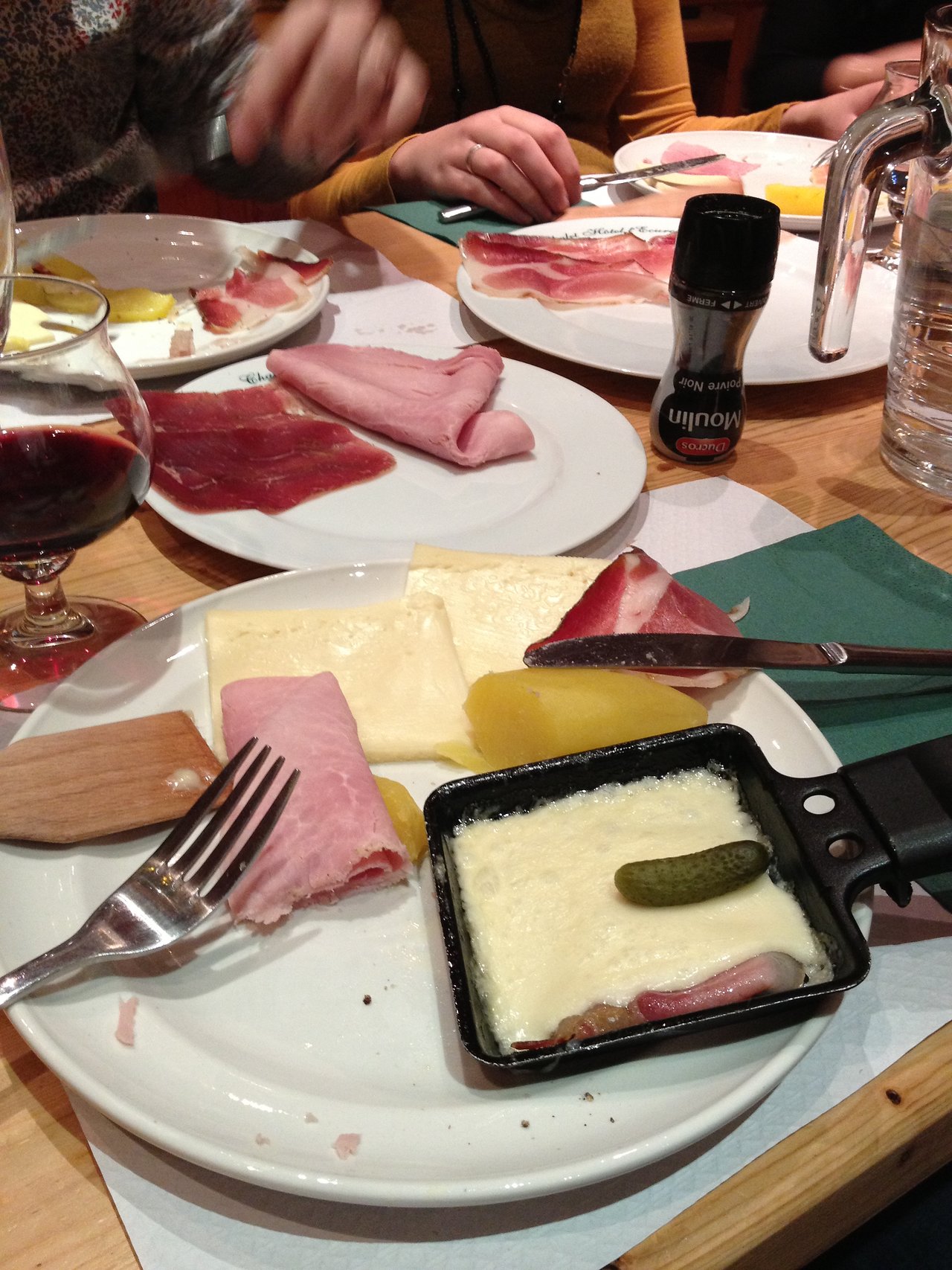 A small pan with melted raclette cheese and a pickle on a plate with ham, potatoes, and utensils.