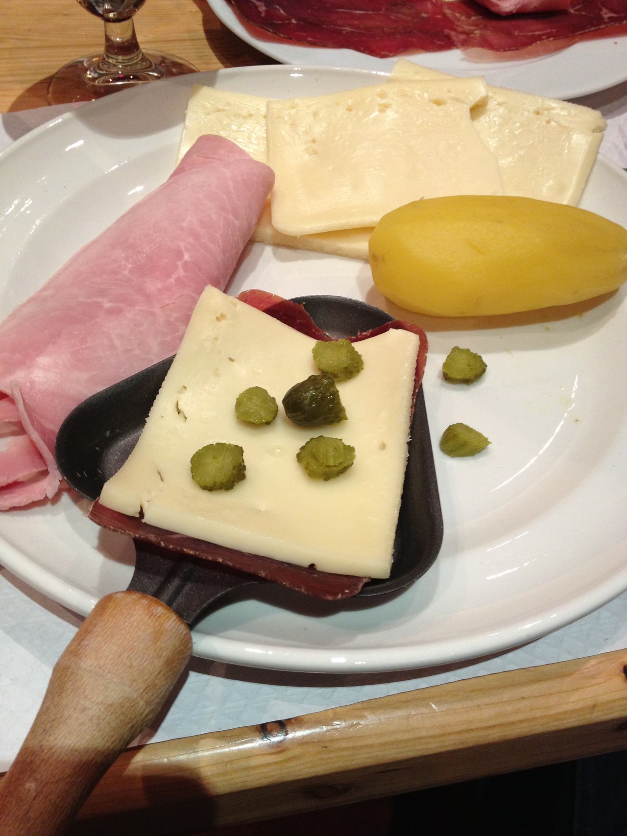A small raclette pan with melted cheese and pickles, placed on a plate with ham, cheese, and a potato.