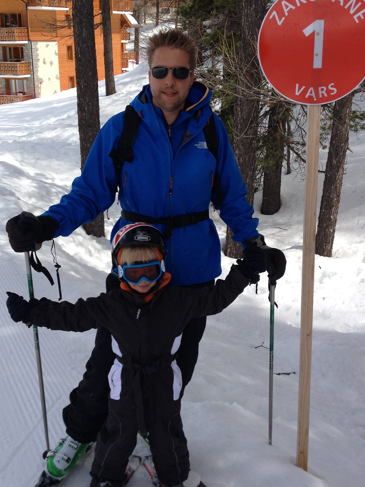 An adult and a child in ski gear stand on a snowy slope near a red Vars ski trail sign.