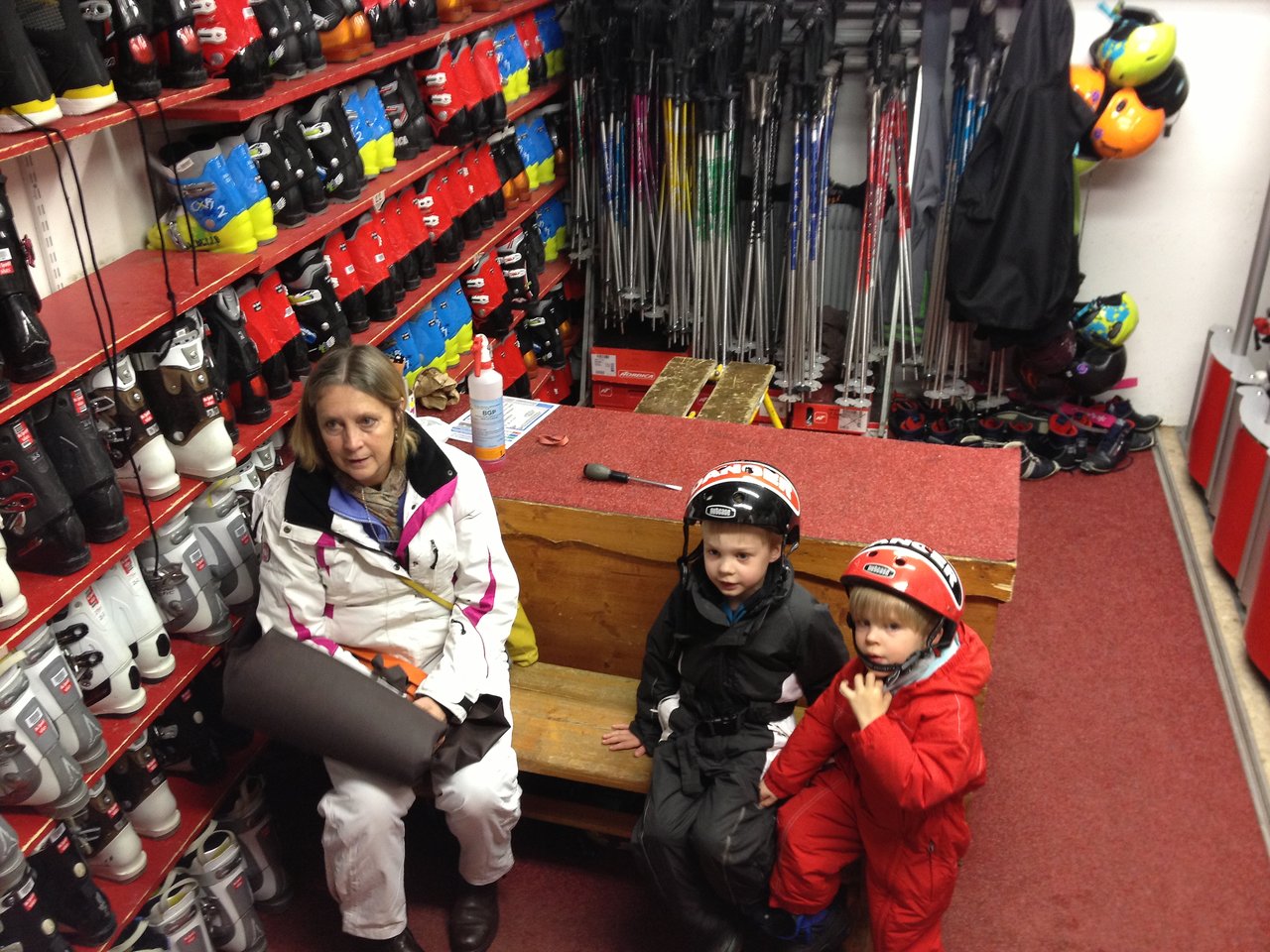 A woman and two children in ski gear sit in a rental shop surrounded by ski boots and equipment.