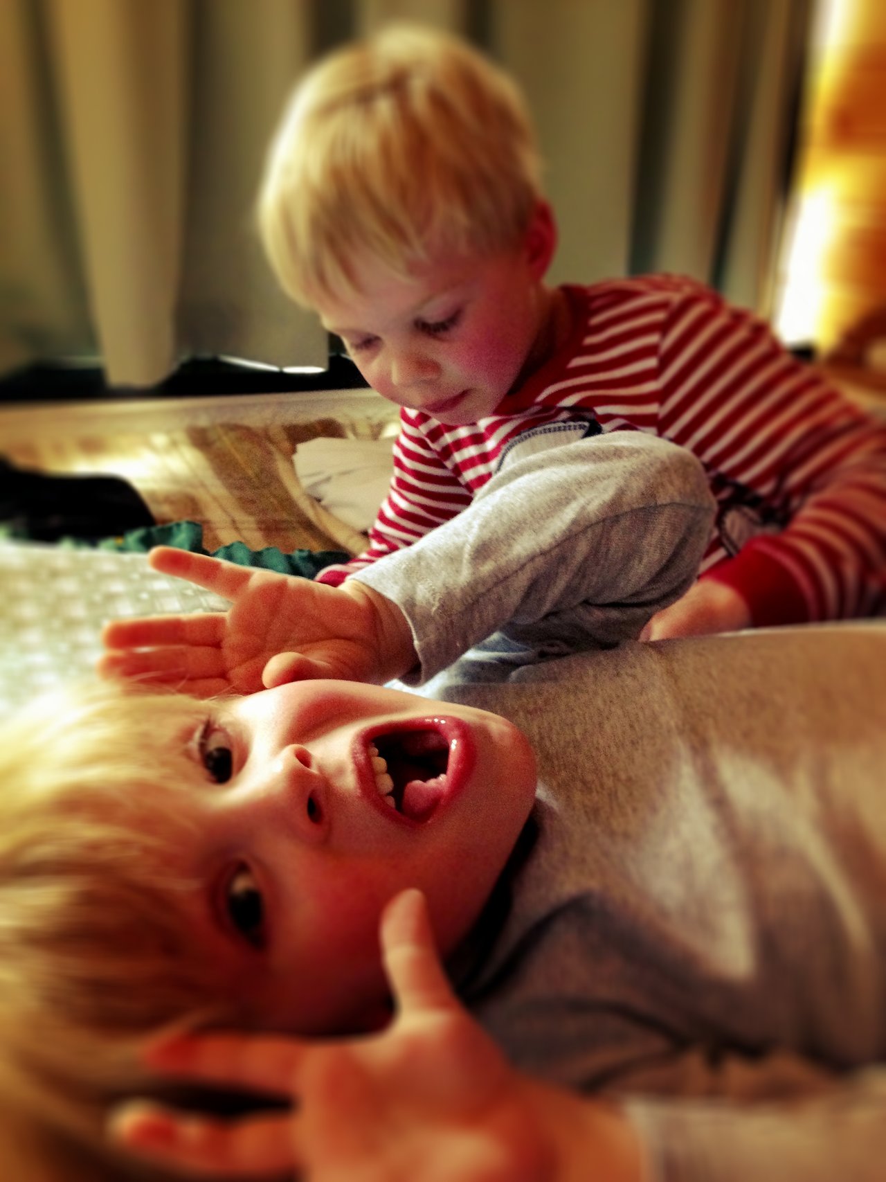 A child in gray lies on their back, making a playful scared face, while another child leans over them.