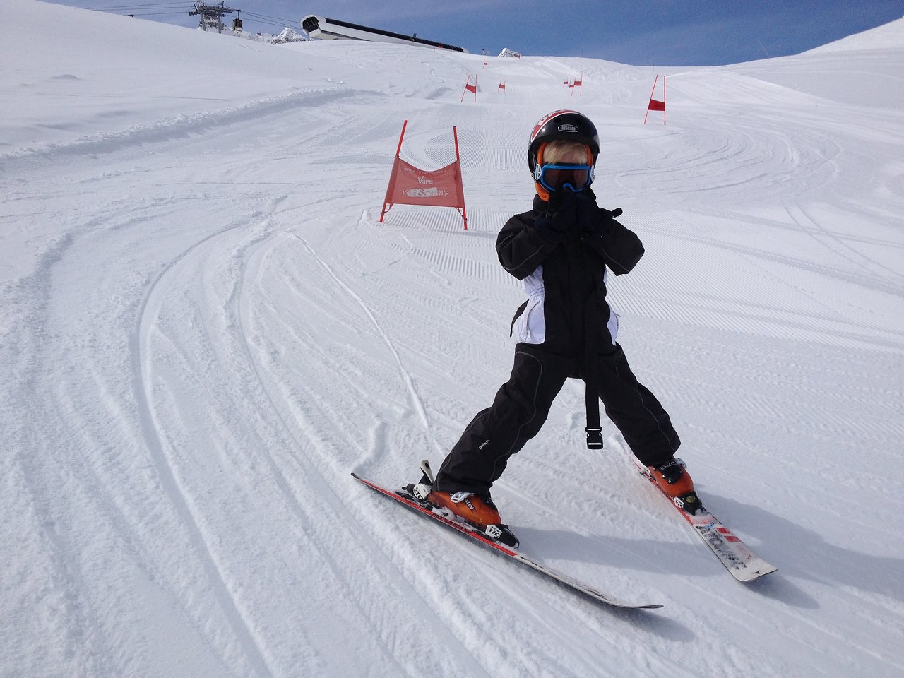 A child in ski gear practices on a snowy slope, wearing a helmet and goggles, with ski gates in the background.
