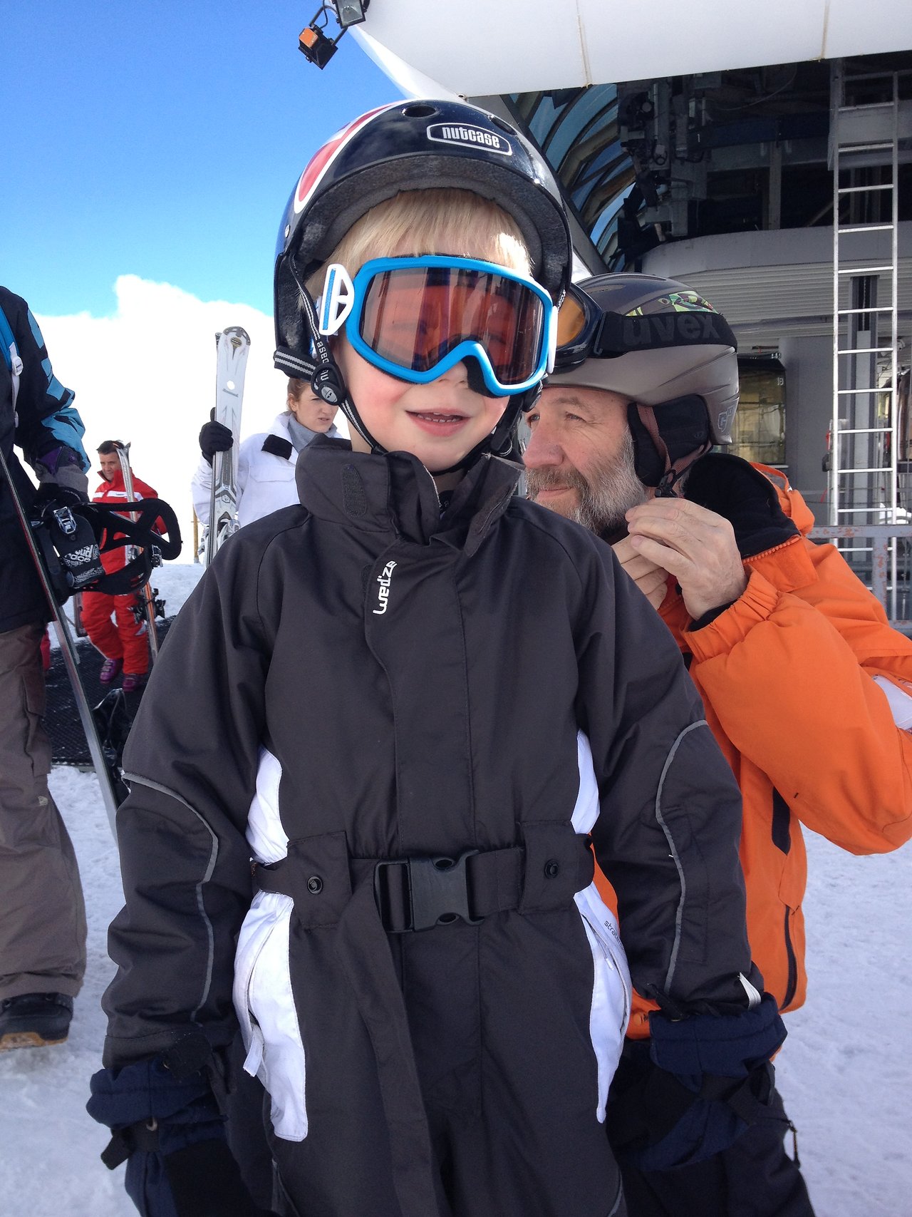 A child in a black ski suit, helmet, and goggles stands on a snowy slope, ready for skiing.