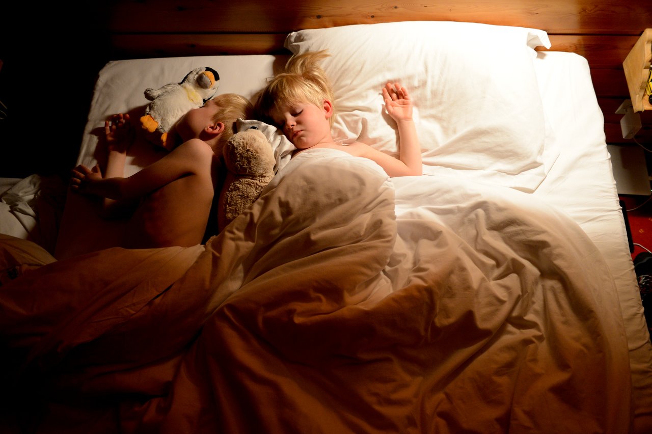 Two children sleep in bed, cuddling stuffed animals under a warm blanket.