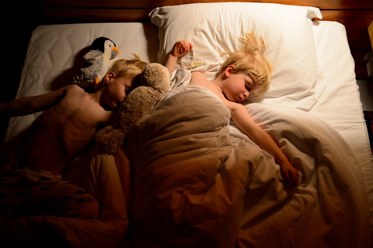 Two children sleep in bed, one partially covered by a blanket, surrounded by stuffed animals.
