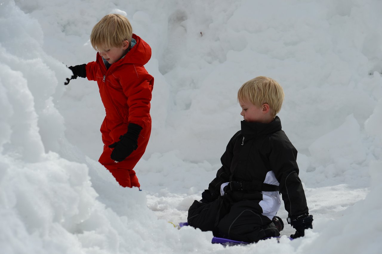 Two children in winter clothes play in the snow, one climbing while the other sits on a sled.