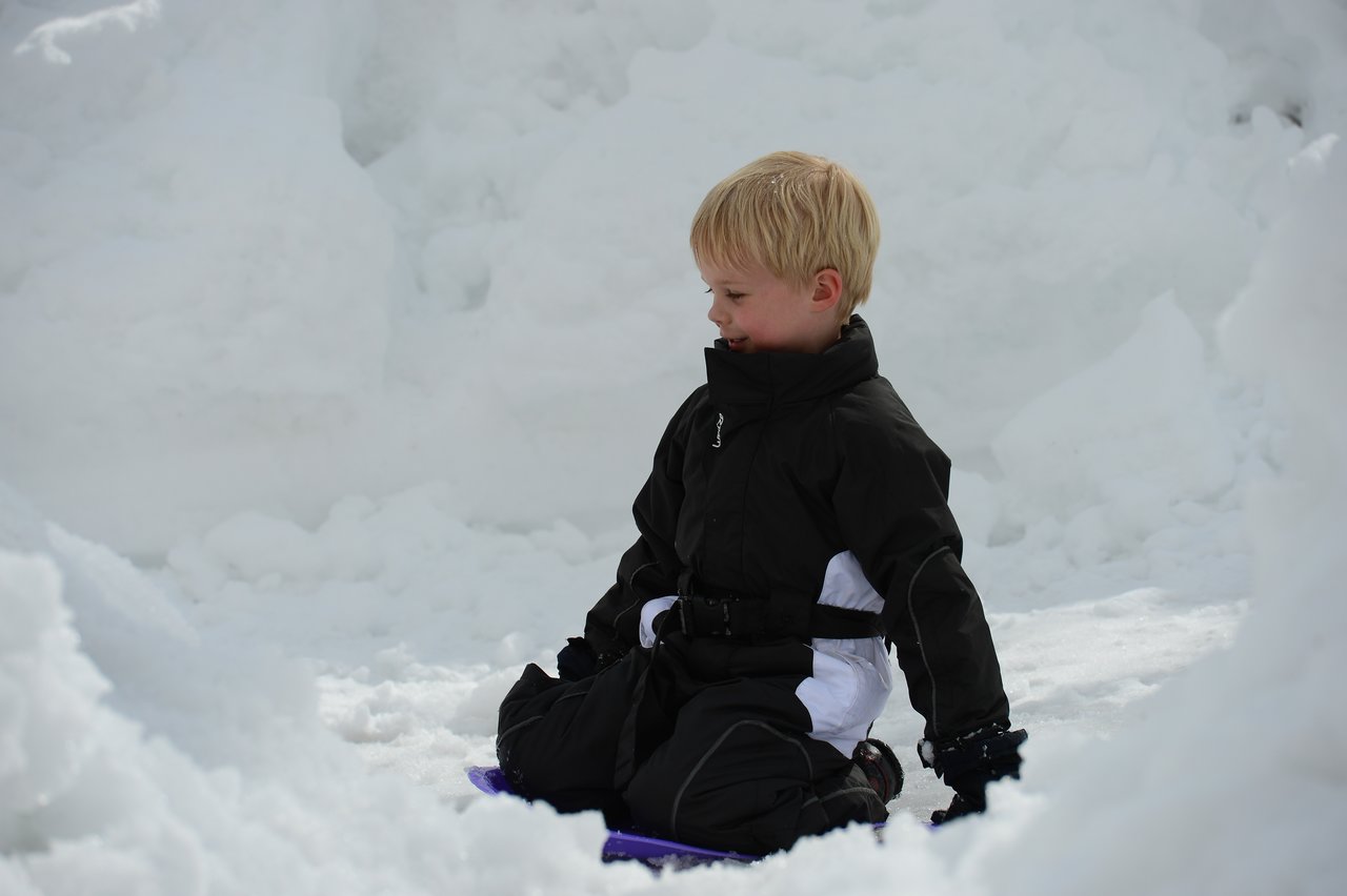 A child in a black snowsuit sits on a small sled in the snow, looking to the side.