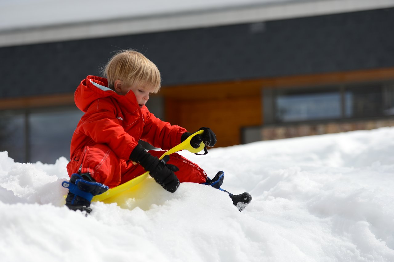 A child in a red snowsuit sits on the snow, holding a yellow sled and preparing to slide.