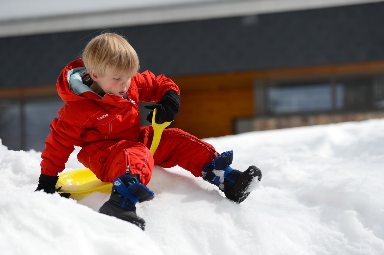 A child in a red snowsuit sits on a snowy slope, holding a yellow sled.