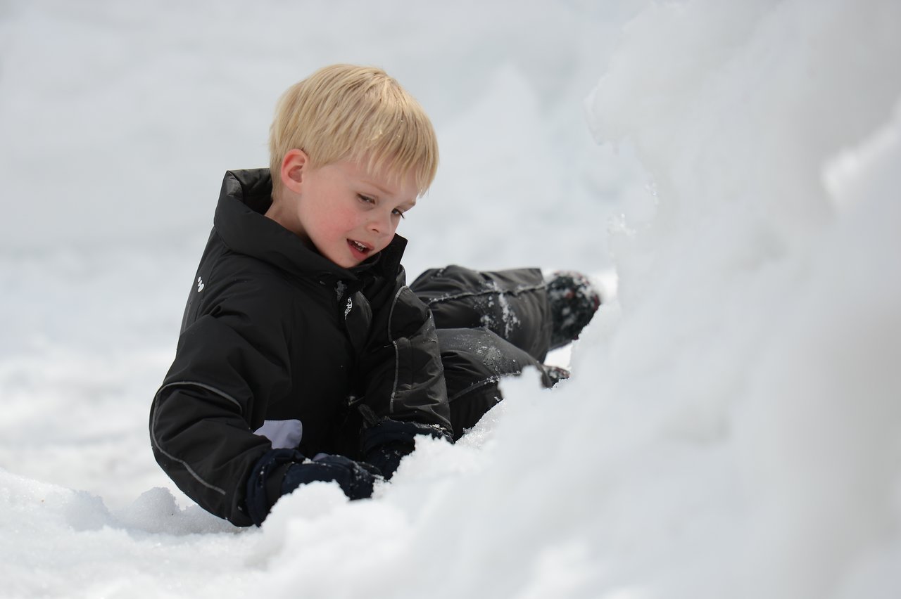 A child in a black winter jacket plays in the snow, smiling while leaning on the ground.