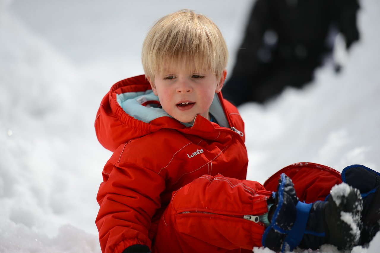 A child in a red snowsuit sits in the snow, wearing winter boots and gloves.
