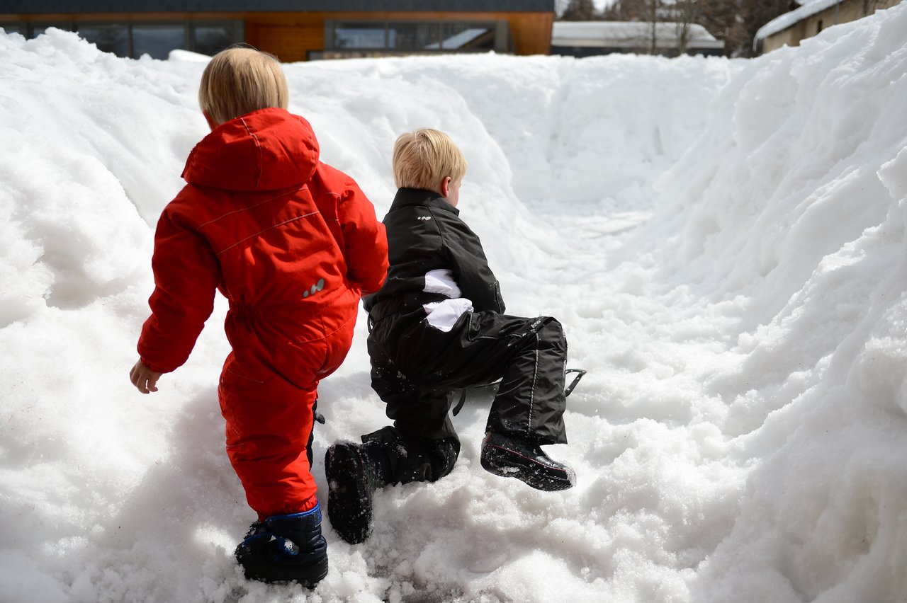 Two children in winter clothes play in deep snow, climbing through a narrow snowy path.