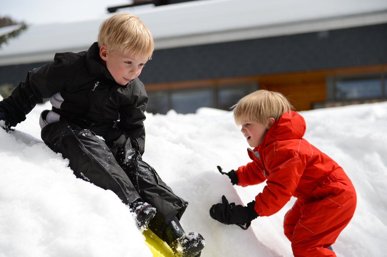 Two children play in the snow; one slides down while the other climbs up, both wearing winter clothes.