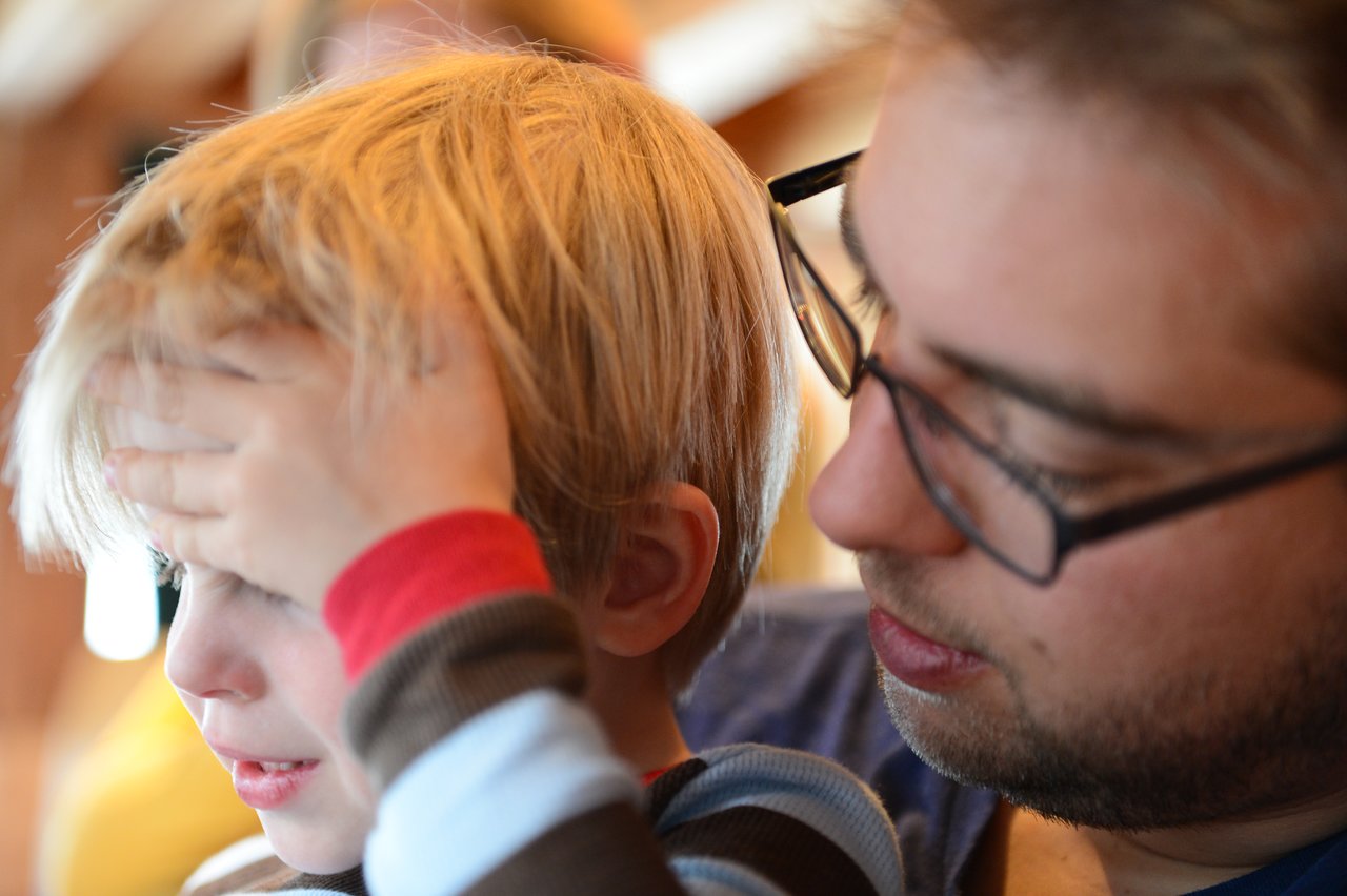 A child covers their face while an adult wearing glasses looks at them closely.