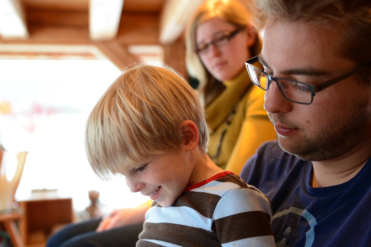 A young boy sits on his father's lap, smiling, while a woman watches in the background.