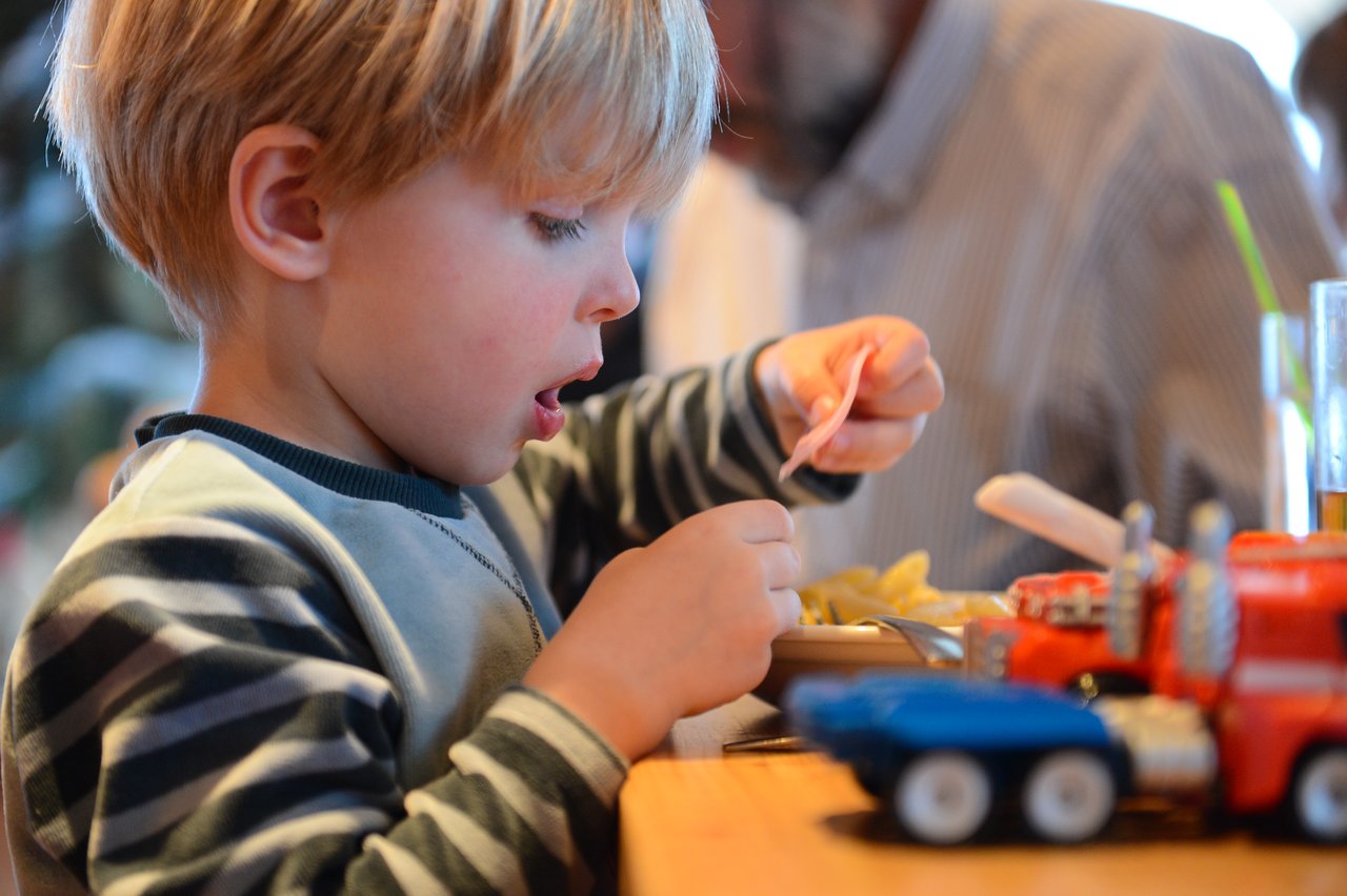 A young child in a striped sweater eats ham with a fork while sitting at a table with toys.