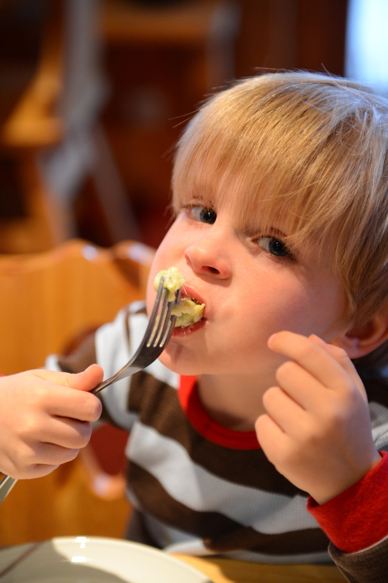 A young child in a striped shirt eats pasta with a fork at a table.