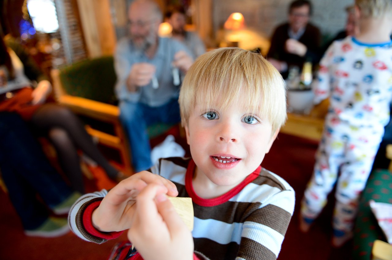 A young child in a striped shirt excitedly holds up an item, with people socializing in the background.