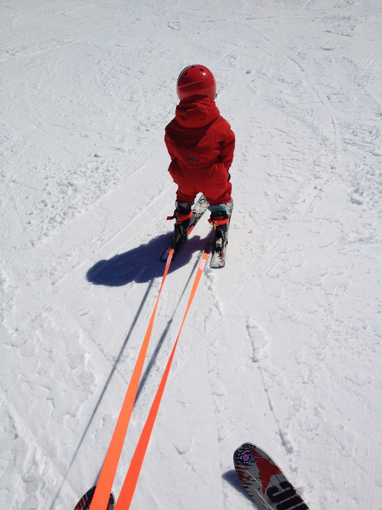 A child in a red ski outfit is skiing with support straps, guided by an adult on a snowy slope.