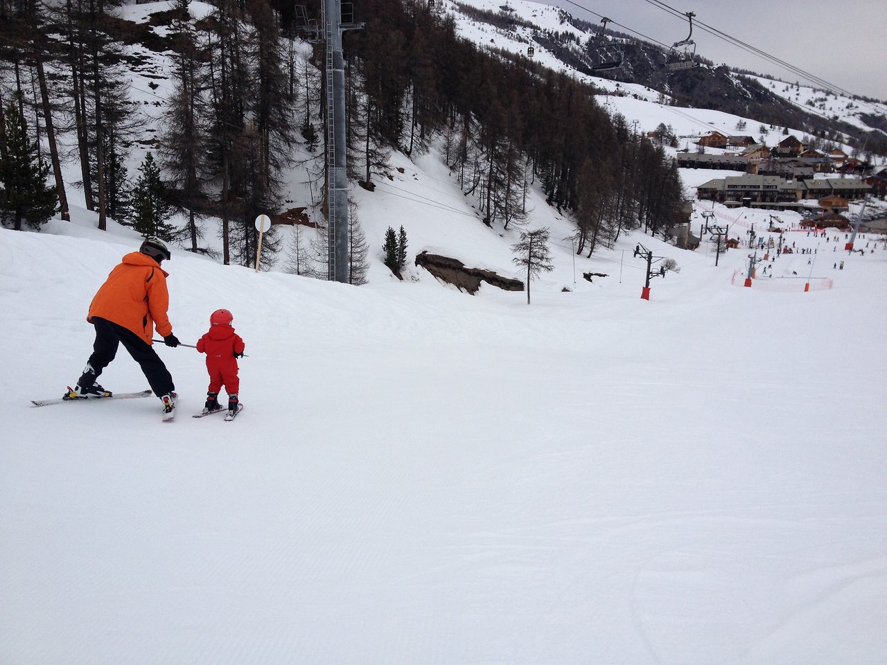 An adult and a child ski together on a snowy slope, with the adult guiding the child from behind.