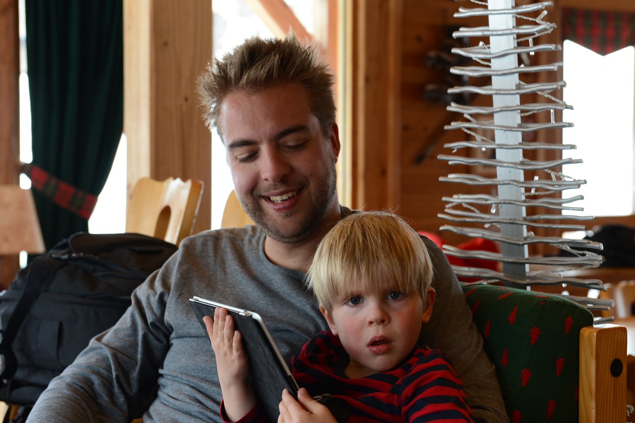 A young child holds an iPad while sitting with a smiling adult in a wooden cabin setting.