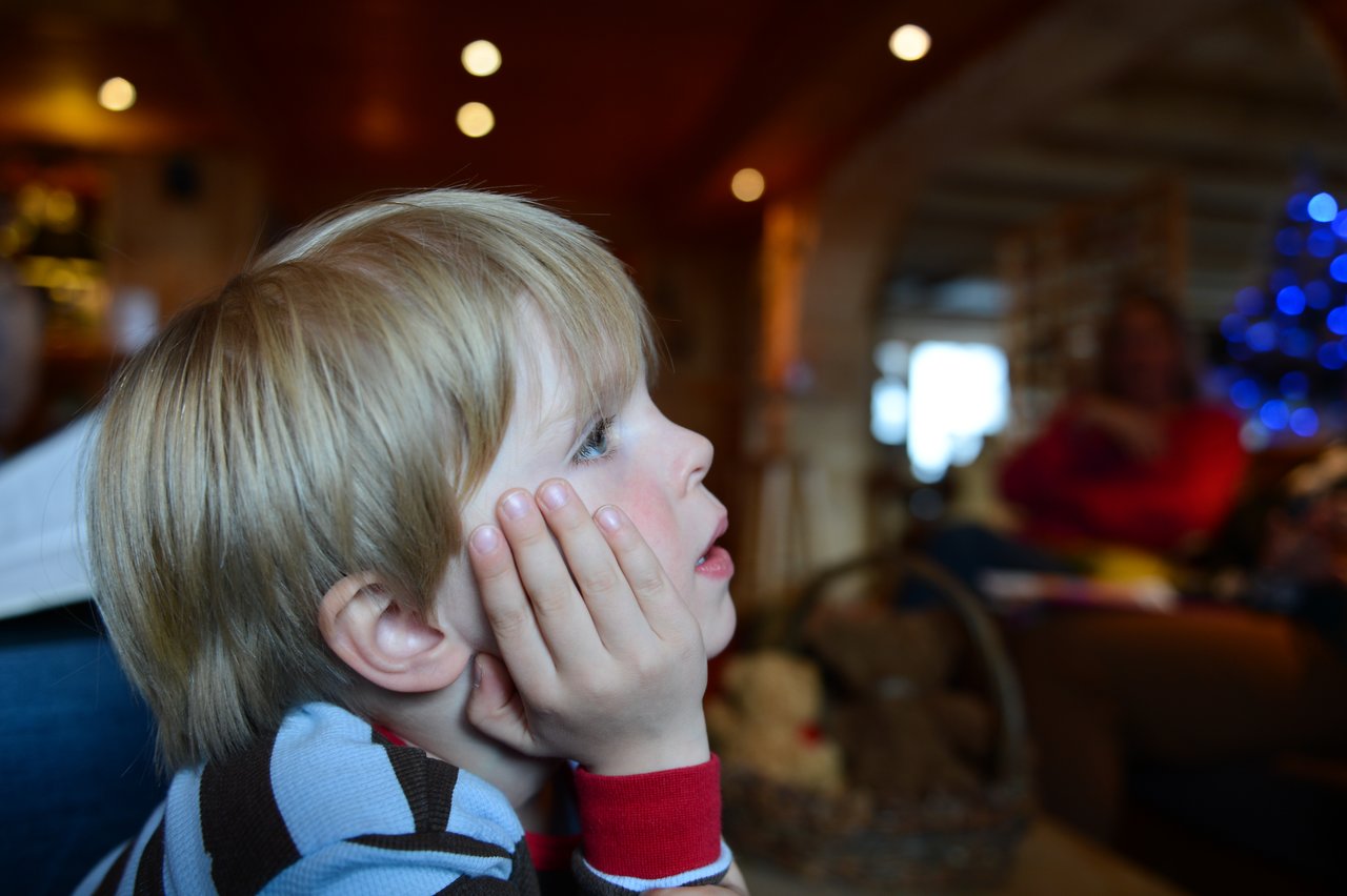 A young child with blonde hair rests their face in their hands, staring intently at something off-camera.