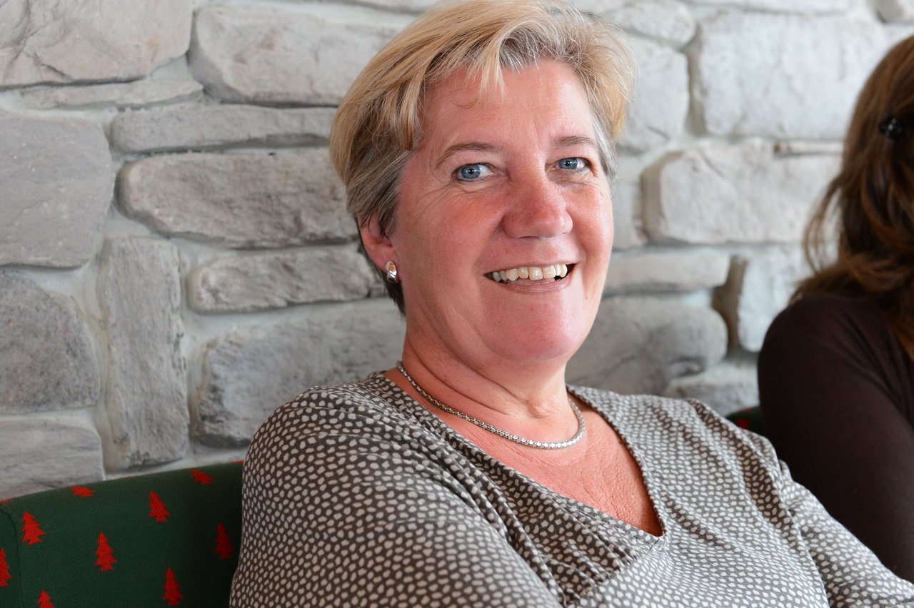 A smiling woman with short blonde hair sits against a stone wall, wearing a patterned top and a necklace.