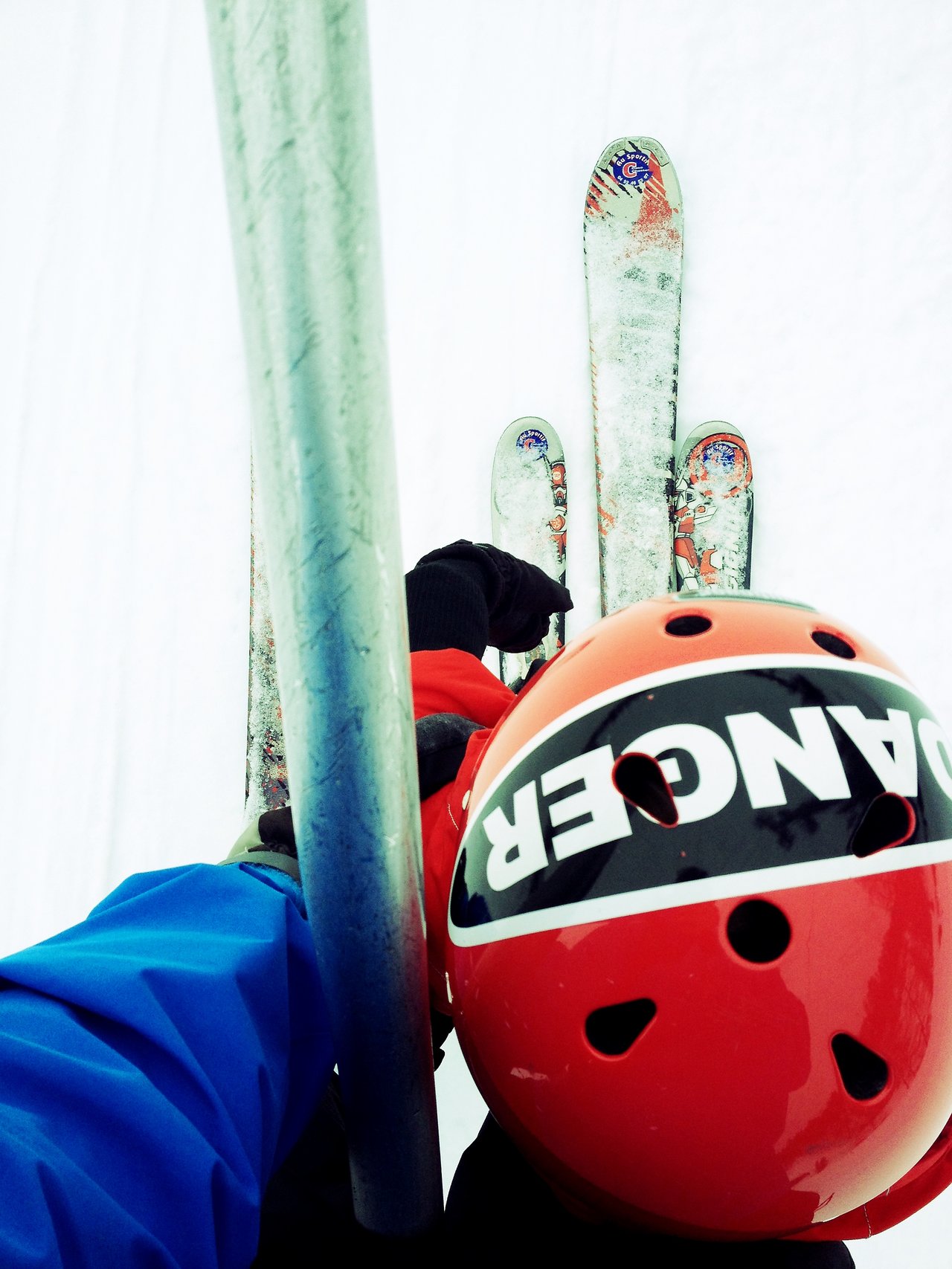Two skiers wearing helmets and winter gear ride a ski lift, holding onto the metal bar for support.