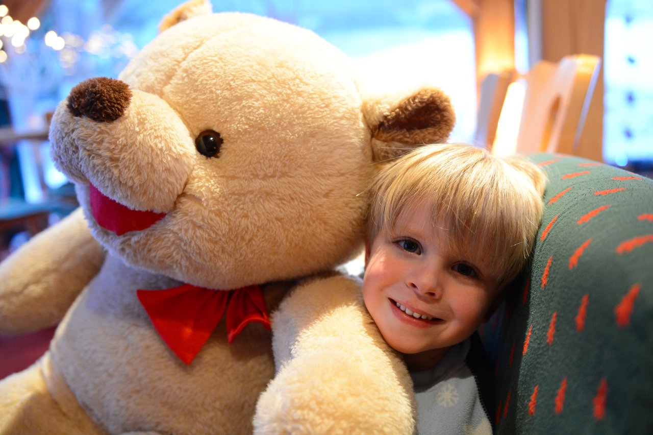 A smiling child sits next to a large teddy bear with a red bow, leaning against a couch.