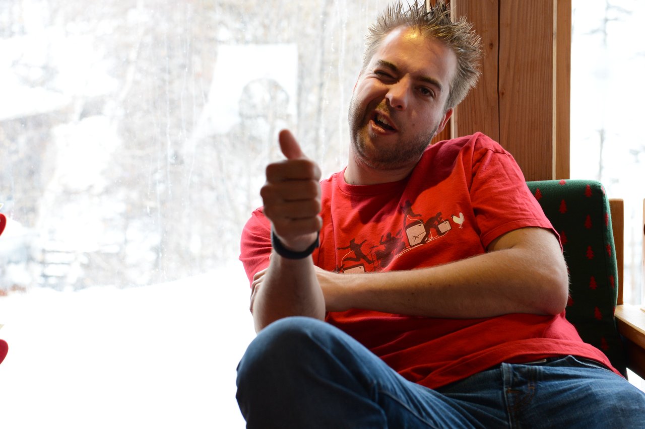 A man in a red shirt sits indoors, giving a thumbs-up and making a playful facial expression.