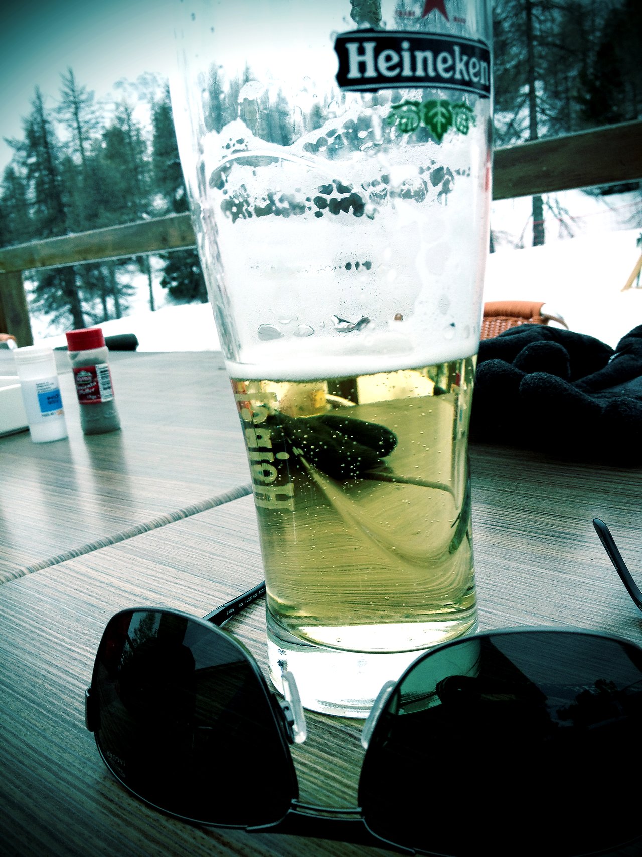 A half-full Heineken beer glass on a wooden table with sunglasses in front, set against a snowy background.