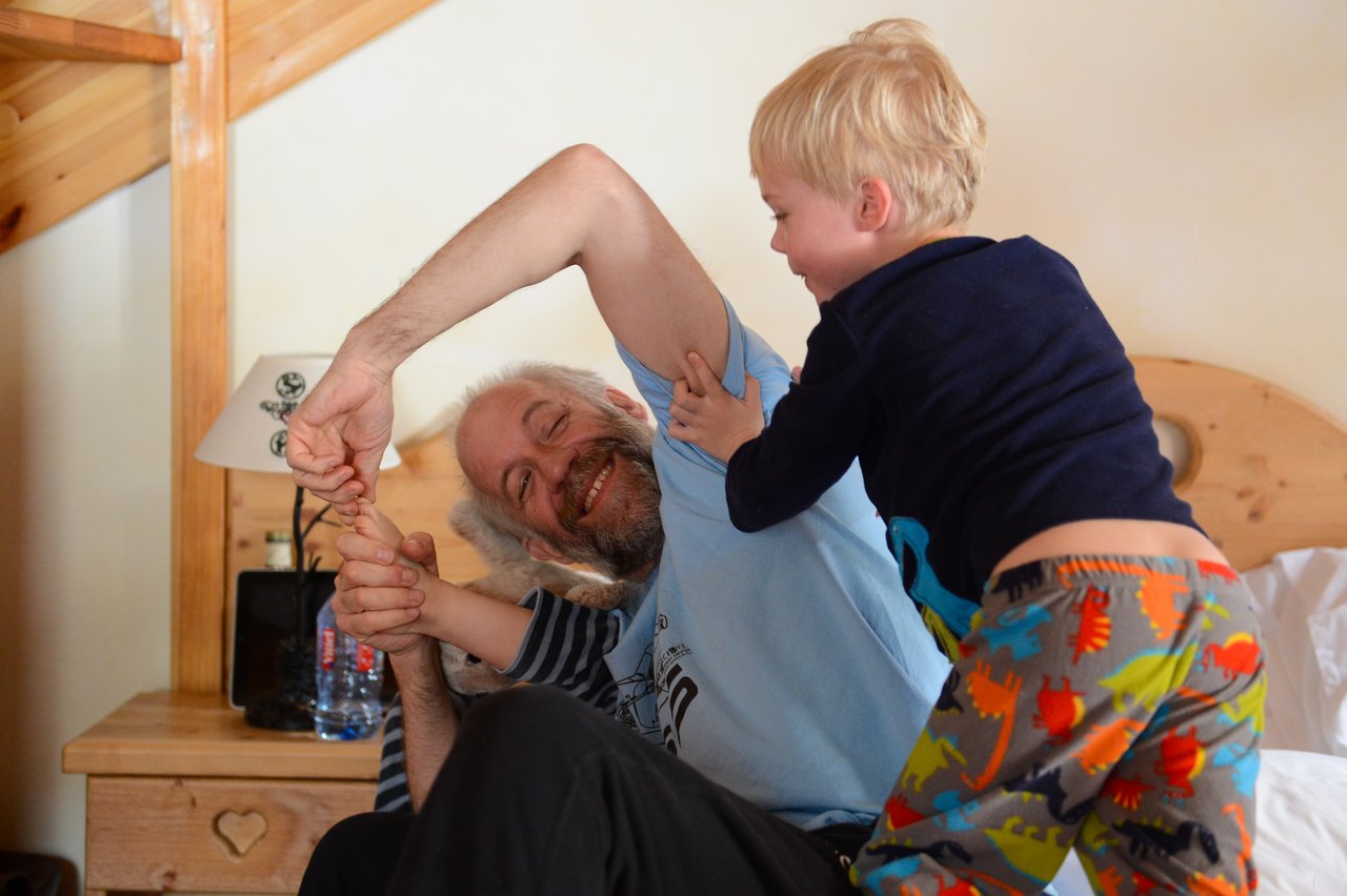 A young child playfully wrestles with an older man on a bed, both smiling and laughing.