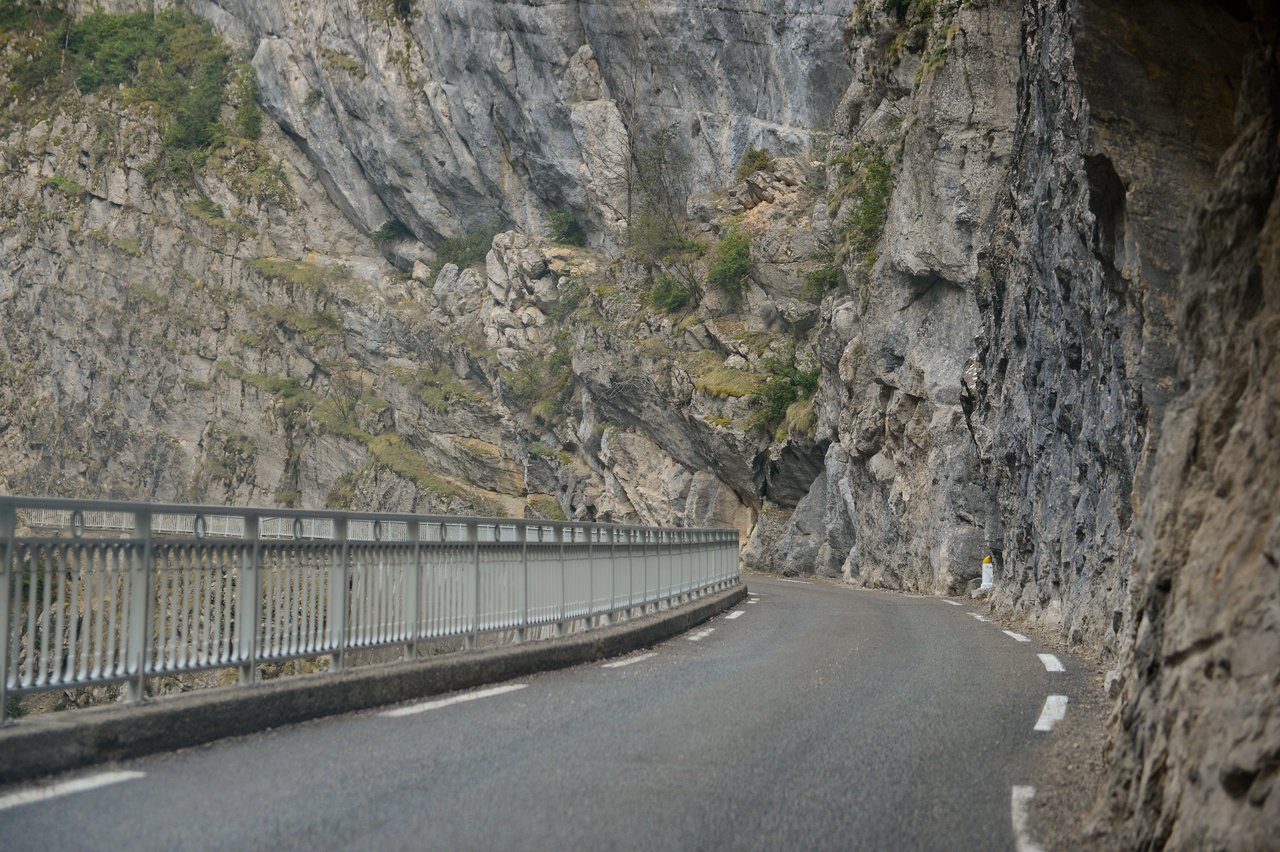 A narrow mountain road curves between a steep rock face and a metal guardrail.