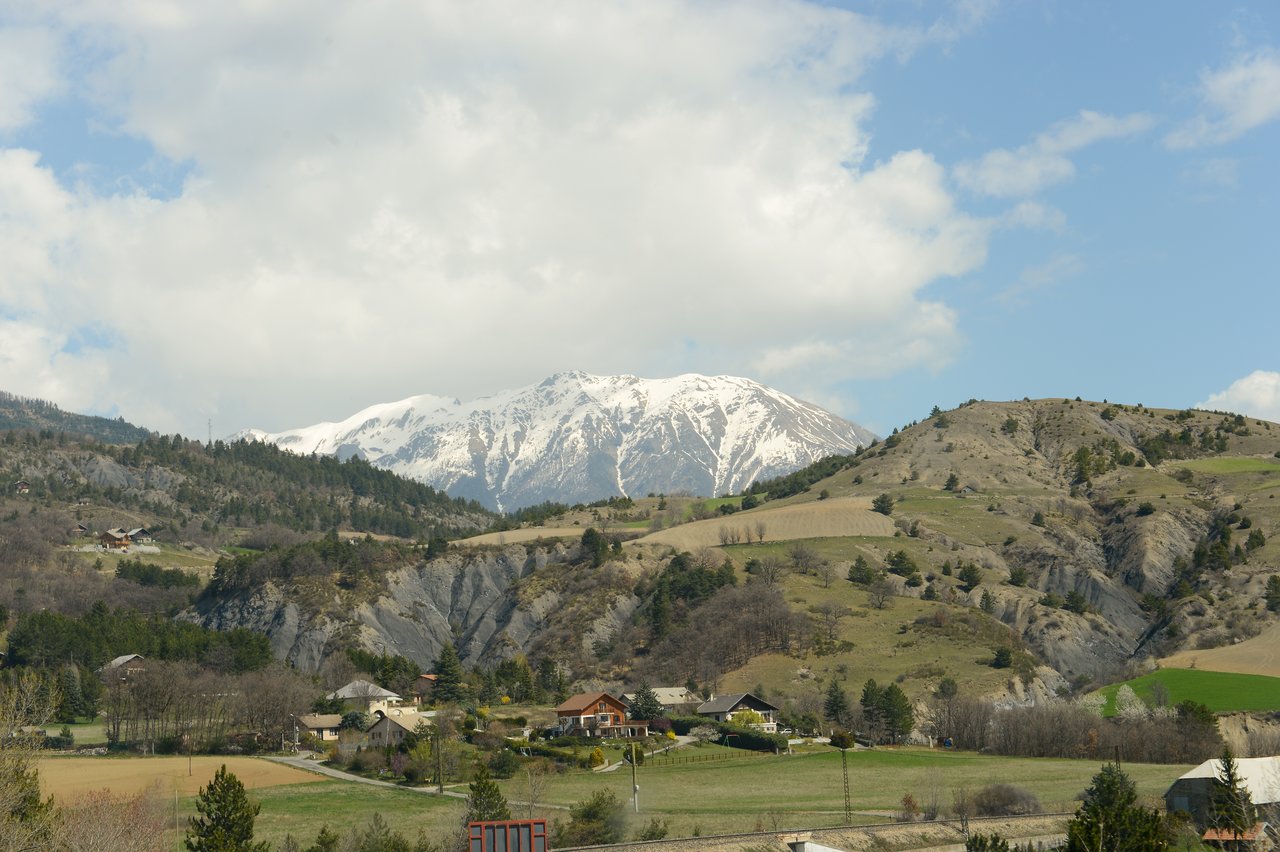 A village with scattered houses sits in a green valley, with a snow-capped mountain in the background.