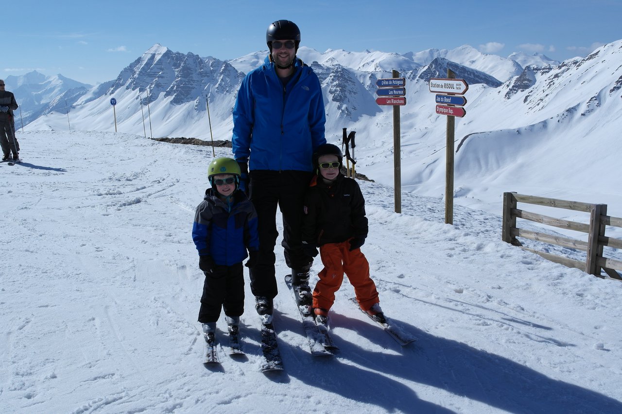 A man and two children in ski gear stand on a snowy mountain with skis on.