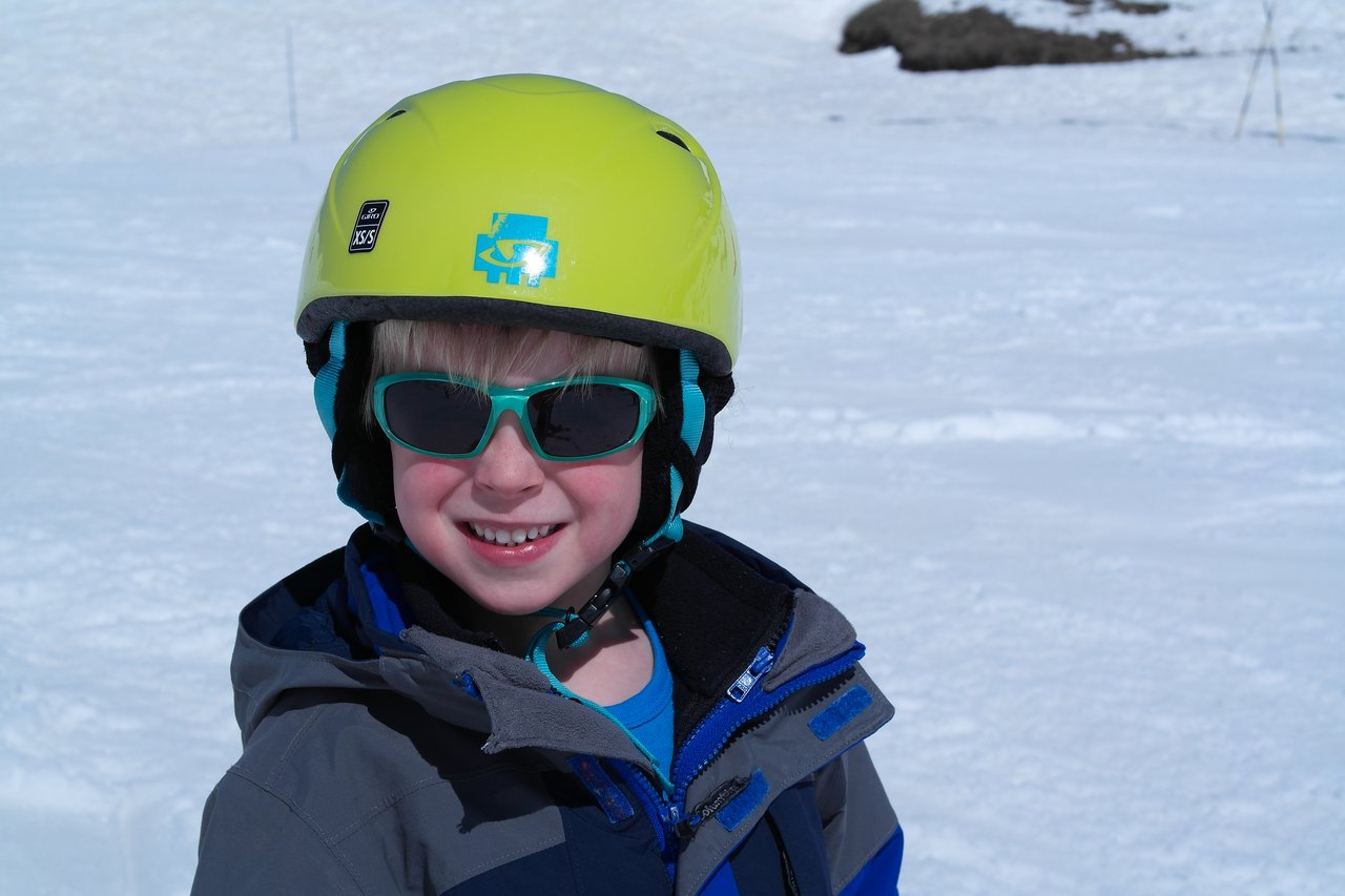 A child wearing a yellow helmet, sunglasses, and a winter jacket smiles while standing on a snowy surface.