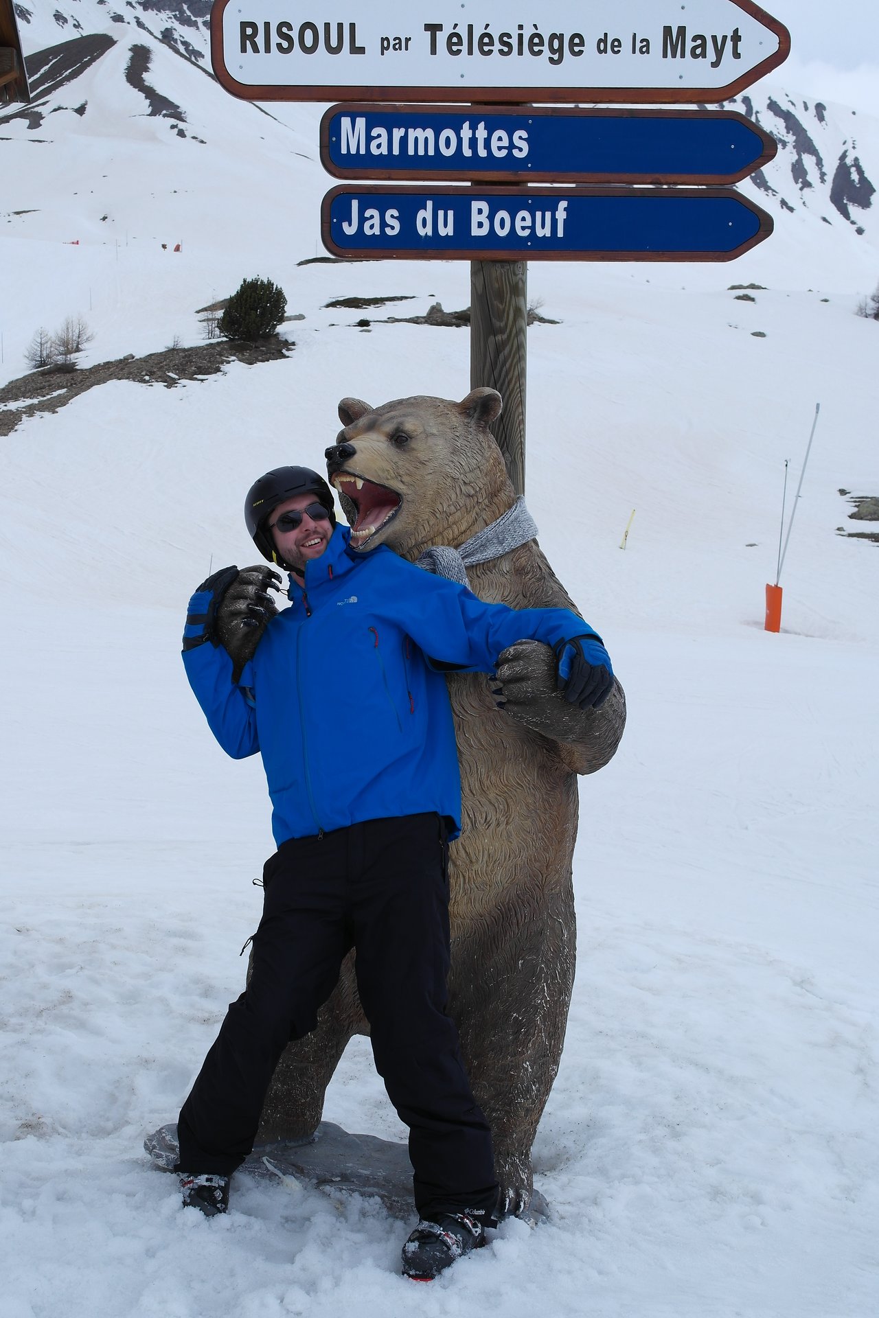 A person in ski gear playfully poses with a bear statue on a snowy slope near directional signs.