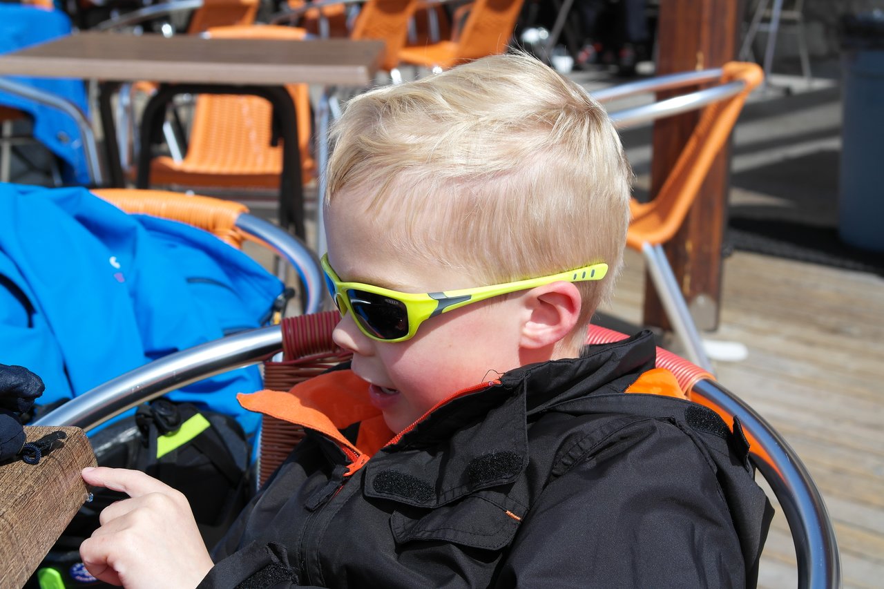 A child wearing sunglasses and a black jacket sits outside, touching a wooden surface with one finger.