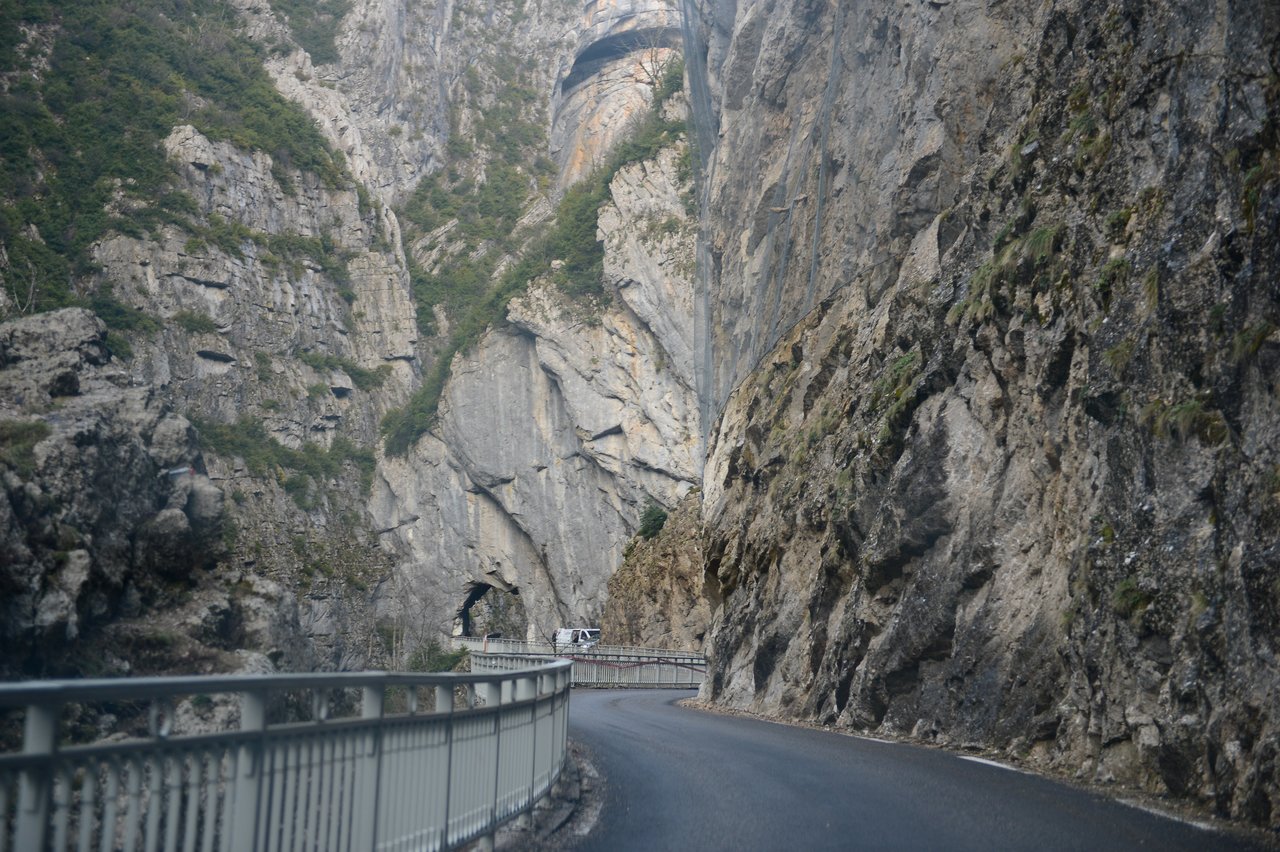 A winding mountain road passes through a narrow rocky gorge, with a vehicle approaching a tunnel in the distance.