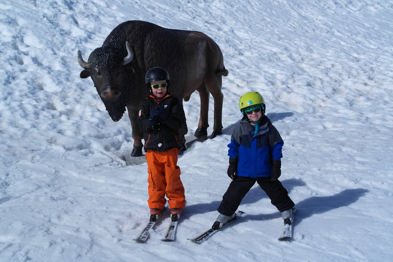 Two children in ski gear pose on the snow in front of a large bison statue.