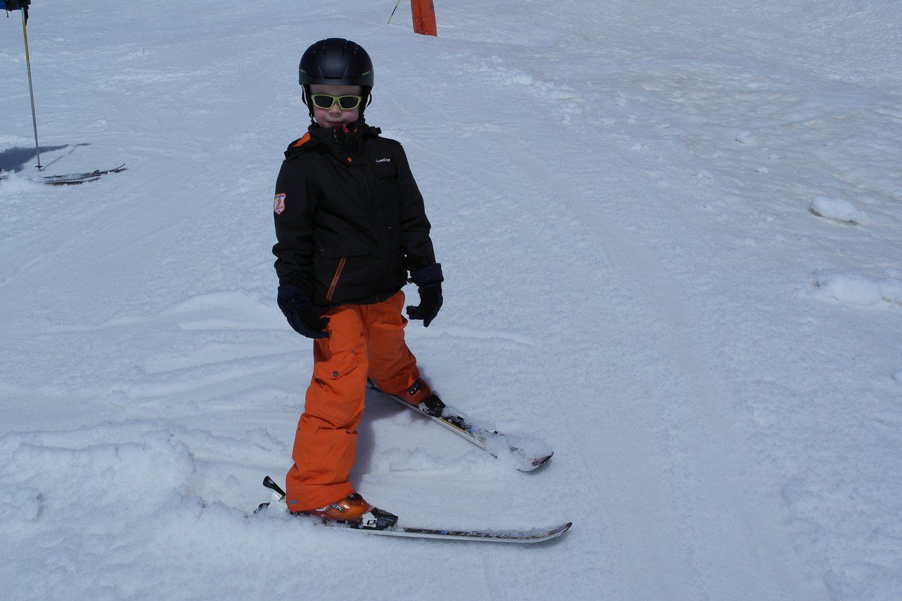 A child wearing a helmet and ski gear stands on skis in the snow, looking at the camera.