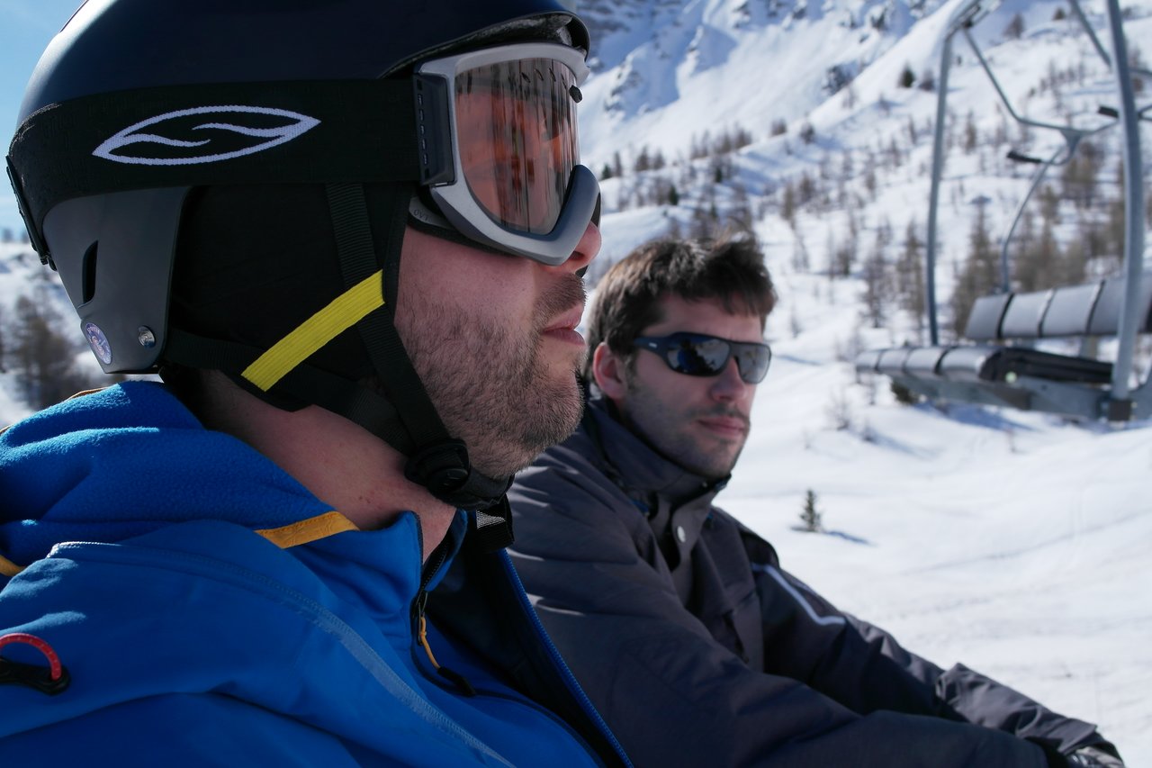 Two men wearing winter gear and sunglasses ride a ski lift in a snowy mountain setting.