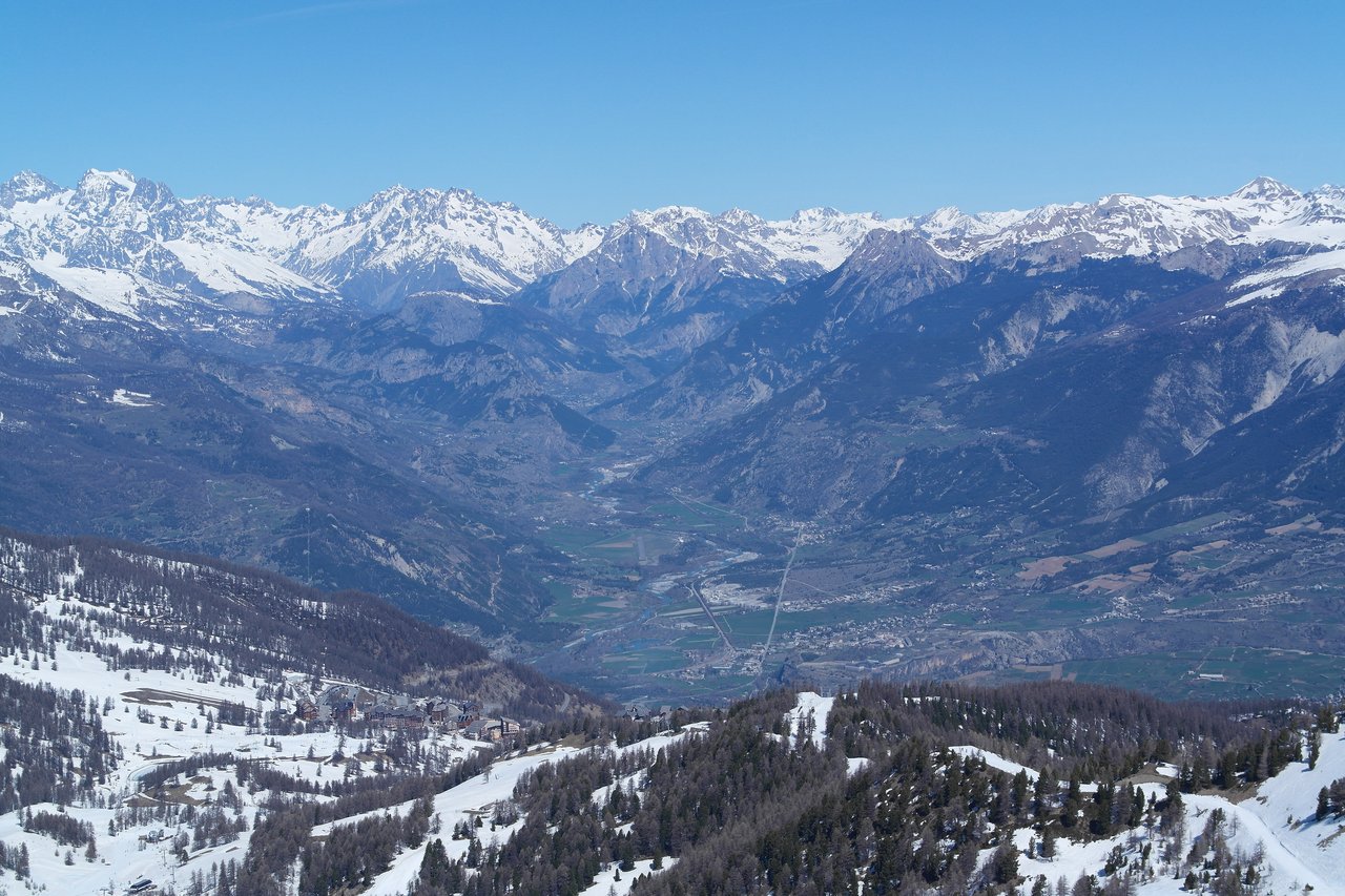 Snow-covered mountains with a valley below, showing a mix of forests, villages, and open land under a clear sky.