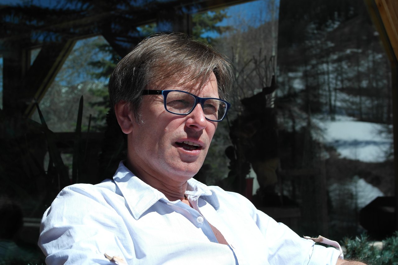 A man wearing glasses and a white shirt sits outdoors, speaking or reacting to something in bright sunlight.