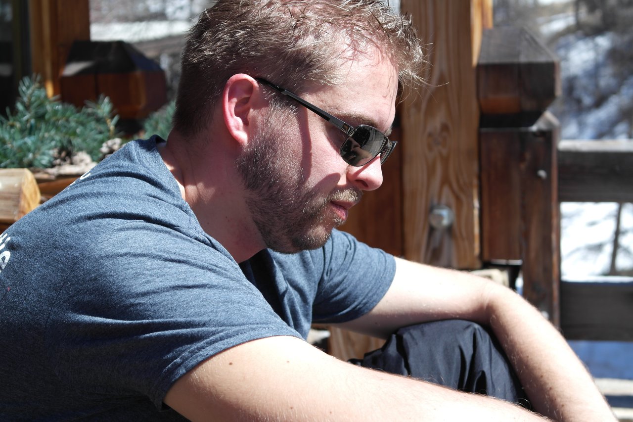 A man wearing sunglasses and a blue shirt sits outside, leaning on his arms in the sunlight.