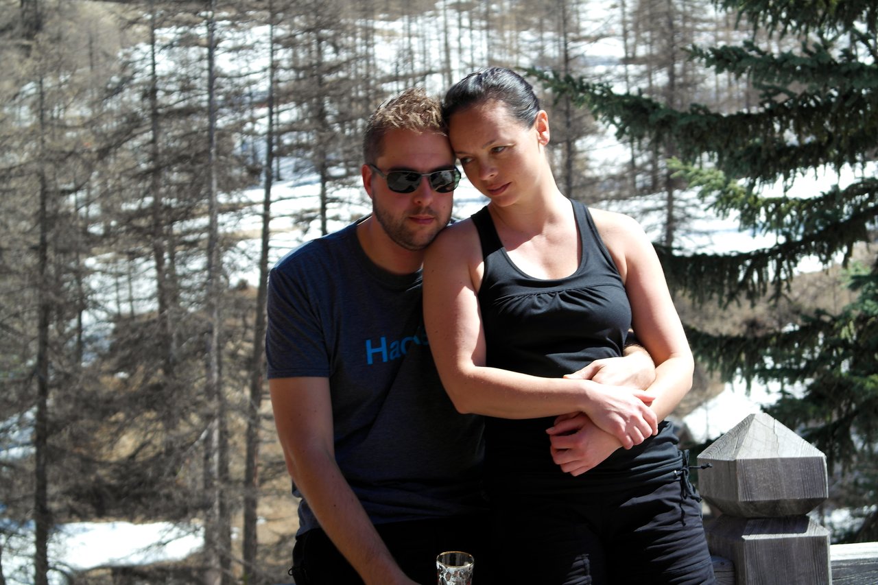 A man wearing sunglasses sits while a woman in black leans against him, wrapping her arms around his.