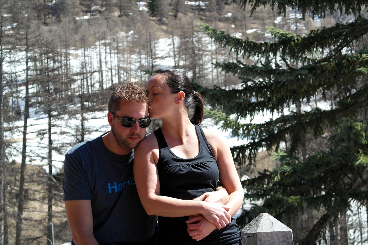 A woman kisses a man's forehead while embracing him outdoors with trees and snow in the background.