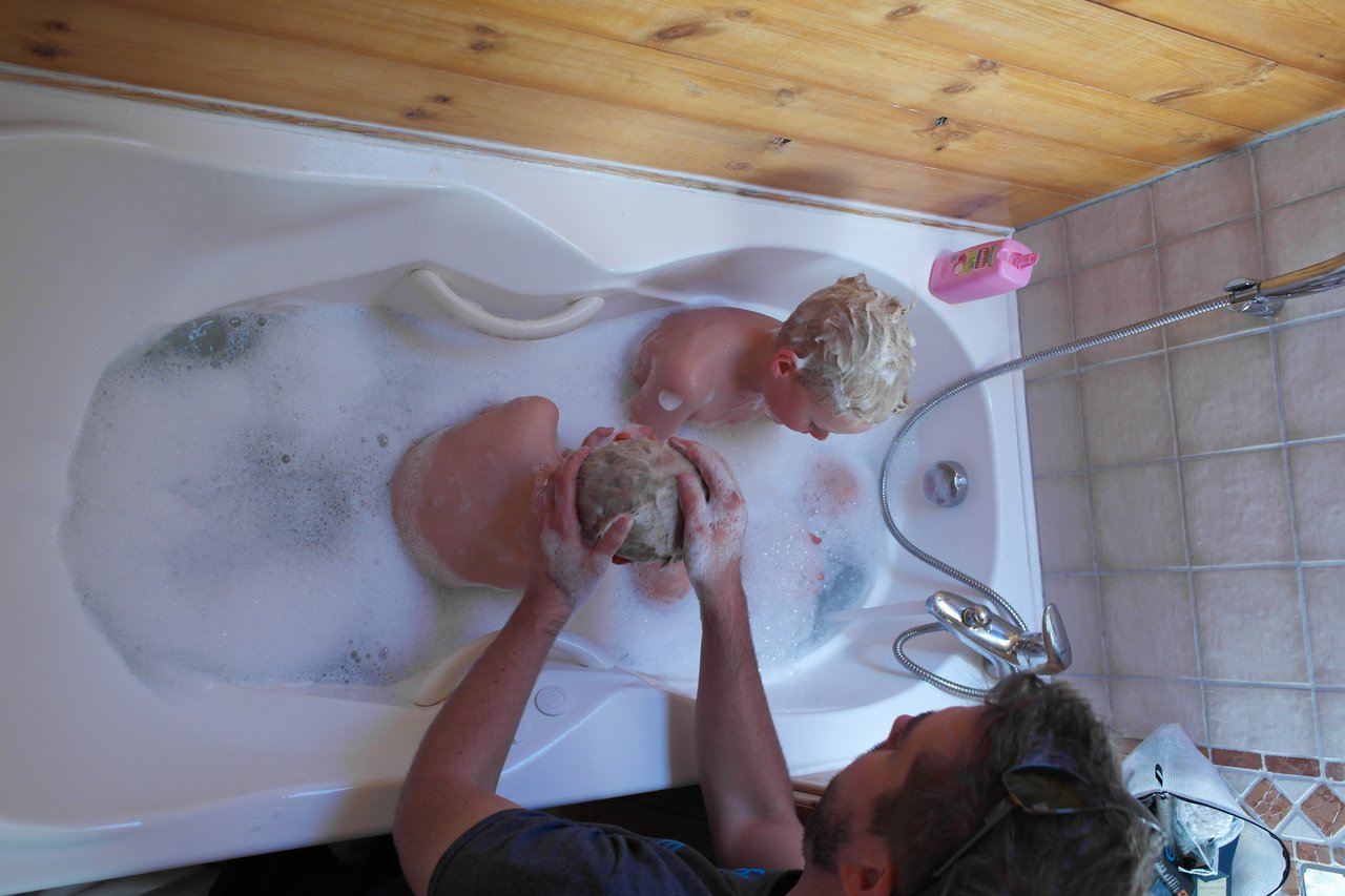 An adult washes a child's hair while two children sit in a bathtub filled with bubbles.