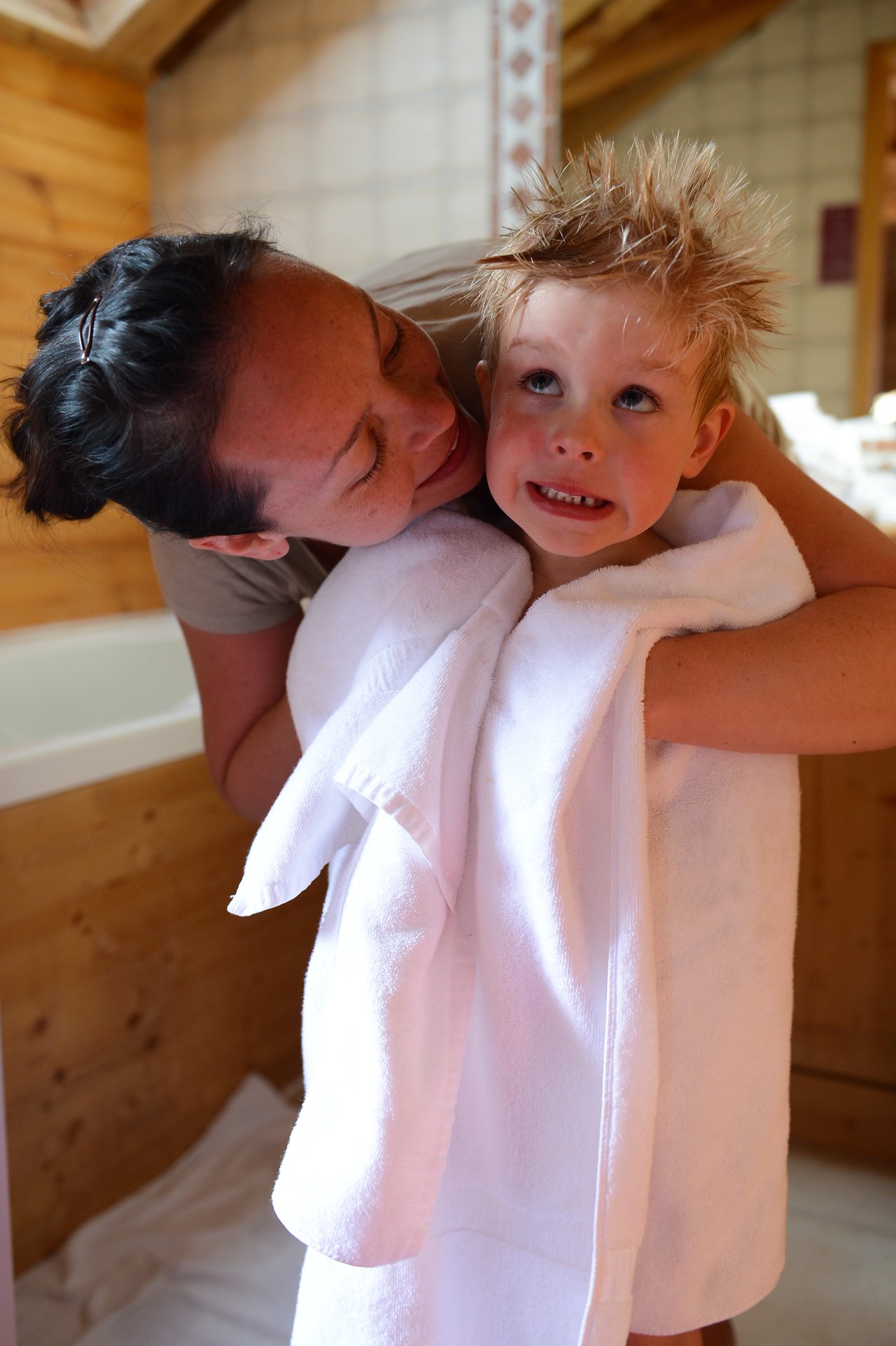 A woman hugs a child wrapped in a towel, while the child makes a playful facial expression.
