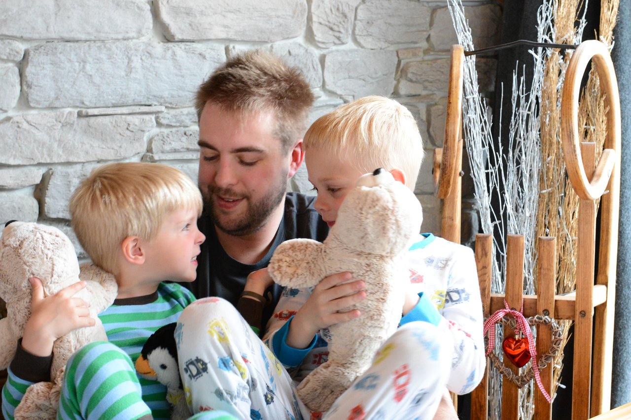 A man sits with two young children in pajamas, both holding stuffed animals and engaging in conversation.